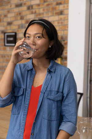 Asian woman wearing denim shirt standing at cafe counter sipping water from clear glass. Casual,...