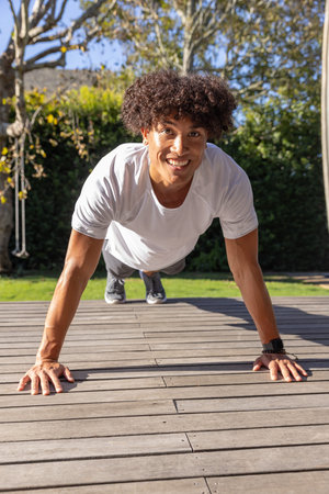 Fitness enthusiast performing push-ups on wooden deck near grass lawn with hedge and swing rope....