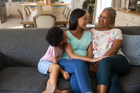 Diverse multigenerational family talking together on grey sofa beside wooden dining table at home....