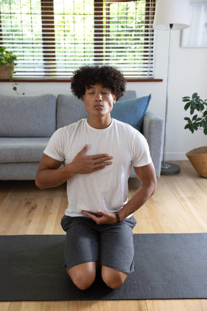 African American man kneeling on black yoga mat in living room practicing breathing exercise. Meditation, relaxation, mindfulness, wellness, interior, home, tranquilityの写真素材