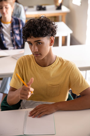 Diverse teenage students raising hand and writing in classroom with yellow pencil and notebook....
