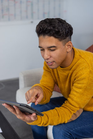 Male in mustard sweater sitting on office sofa with rust cushion tapping tablet near planning board....