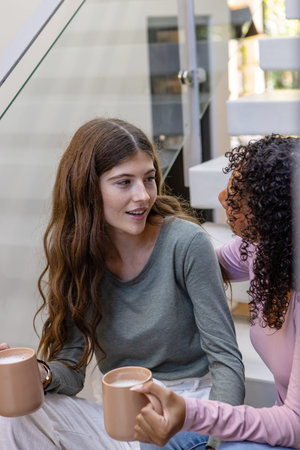 Diverse female friends holding mugs and chatting on modern staircase landing at home. Friendship,...