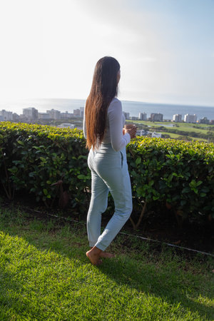 Indian woman standing barefoot on terrace behind manicured hedge holding mug and gazing at skyline....