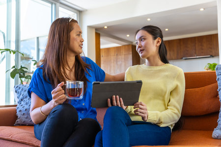 Asian mother and daughter sitting on orange couch in living room holding glass mug and tablet. Cozy,...