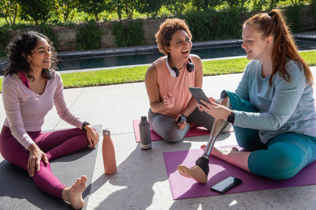 Diverse female friends sitting on yoga mats beside pool sharing tablet with visible prosthetic leg....