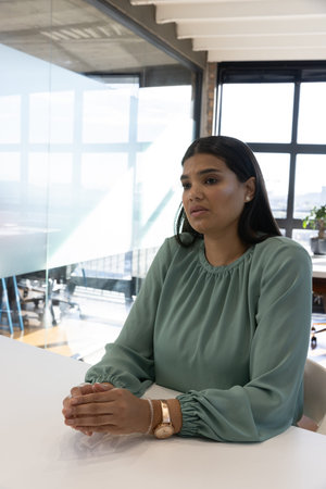 Mid adult woman sitting at white conference table wearing sage blouse near glass wall, copy space....