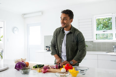 Asian man standing at white countertop in modern kitchen chopping red onion with chef's knife....