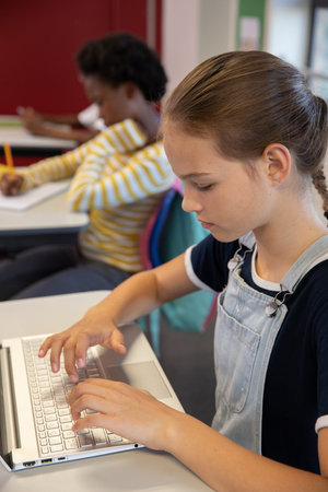 Diverse female classmates typing on laptop and writing on paper at desks in classroom, copy space....