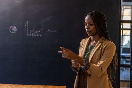 African American woman in tan blazer presenting chalk diagrams on chalkboard, copy space. Educator,...