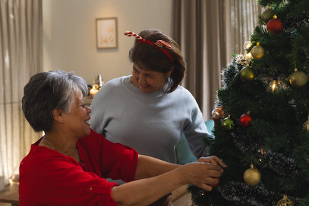 Asian mother and daughter placing gold bauble on Christmas tree with string lights in living room. Festive, celebration, warmth, family, tradition, cozy, holidayの写真素材