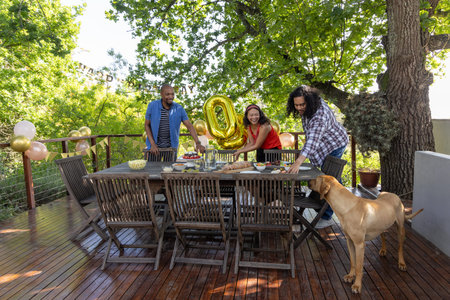 Diverse friends arranging party setup on wooden deck with dining table gold '0' balloon and dog....