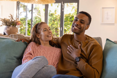 Diverse couple sitting on sofa with teal cushions beside vase at home near glass doors smiling....
