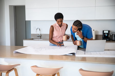 Diverse couple examining blueprints at kitchen island, pointing details and using smartphone....