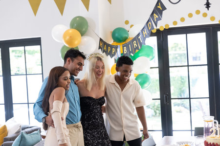 Diverse friends standing at home dining table under Happy Birthday banner with balloons and cake....