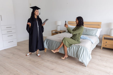 Diverse female sisters practicing graduation speech in gown using tablet in bedroom, copy space....