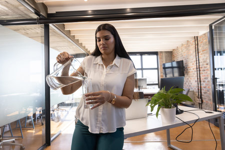Woman in business casual pouring water from glass pitcher into glass at office desk with laptop....