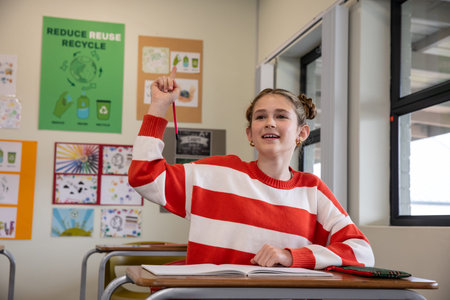 Female child student raising hand at desk in classroom with red pencil over open notebook....