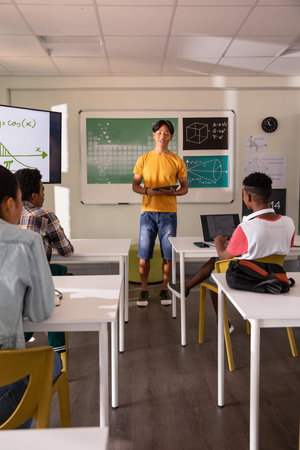 Teenage Asian boy standing at front holding tablet and presenting formulas to classmates in class....