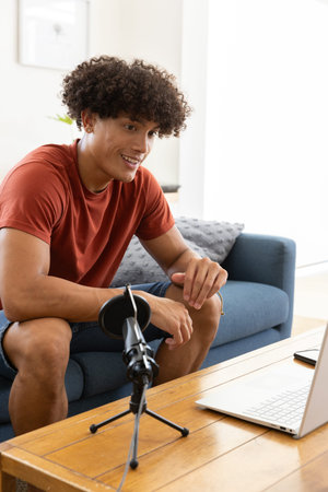 African American man sitting on blue sofa at home recording podcast with laptop, pop filter mic....
