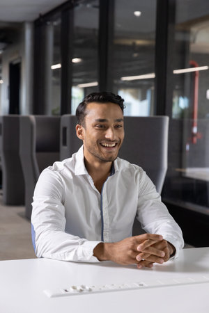 Asian man sitting at white conference table near gray pods in office lounge wearing button-up shirt....