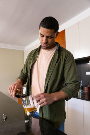 African American man pouring beer into clear glass mug at kitchen counter with bottle opener....
