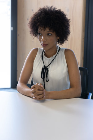 African American woman sitting and clasping hands at white table near large window wearing blouse....