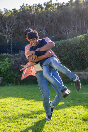 Asian father and son playing in sunny backyard with brick border, father lifting son joyfully. Joy,...