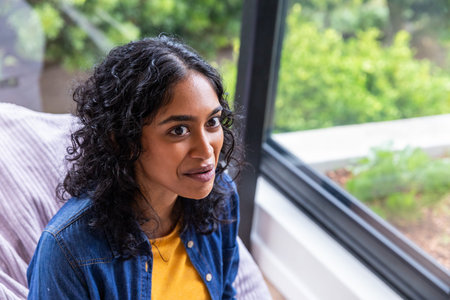 Asian woman sitting on sofa by window at home, in denim shirt beside throw blanket. Cozy, elegance,...