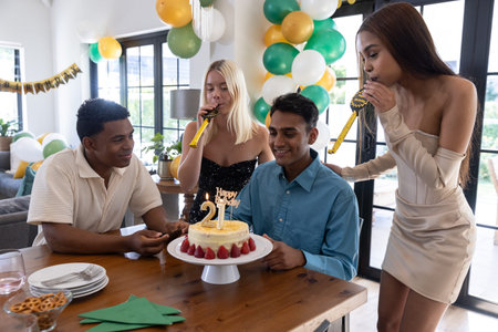 Diverse friends in party attire gathering around table at home celebrating birthday cake, balloons. Festivities, camaraderie, celebration, joyful, decor, leisure, gatheringの写真素材