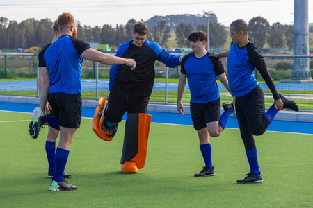 Diverse male hockey players linking arms, stretching quadriceps on turf wearing orange goalie pads. Athletes, teamwork, sportswear, outdoor, dynamic, fitness, competitionの写真素材