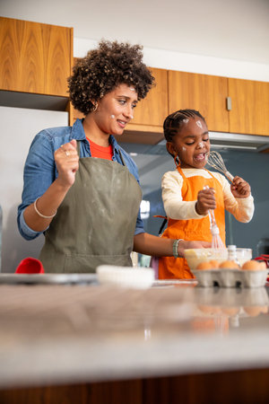 African American mother and daughter in aprons whisking batter with whisks on countertop in kitchen....