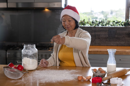 Senior Asian woman wearing Santa hat sprinkling flour beside glass jar on countertop in kitchen....