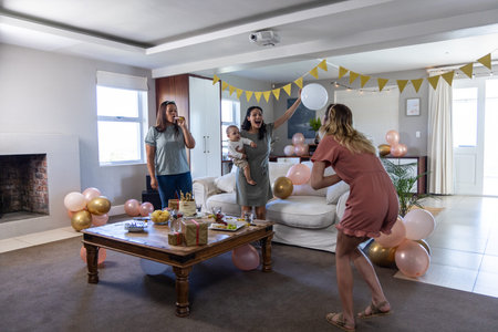 Diverse women and baby boy playing around coffee table in living room with cake and balloons....