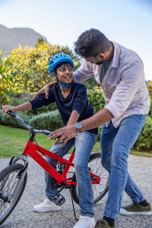 Asian father and son balancing on red bicycle at garden patio wearing blue helmet, watch, sneakers....