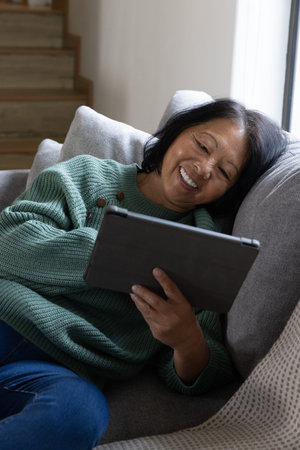 Senior Asian woman reclining on gray couch by stairs holding tablet and smiling with pillows....