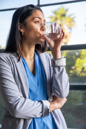Female professional standing by large window sipping water from glass with palm greenery. Bright,...