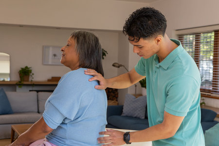 Senior Indian woman receiving back massage from male caregiver in living room with sofa and plants....