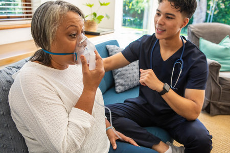 Senior African American patient and Asian nurse in scrubs sitting on sofa using oxygen mask....