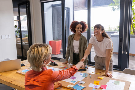 Diverse female coworkers in business attire shaking hands around table at office with sticky notes....
