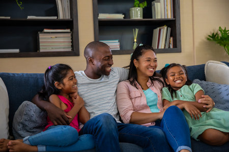 Diverse family sitting on cushioned sofa in living room, surrounded by decorative plants and books....