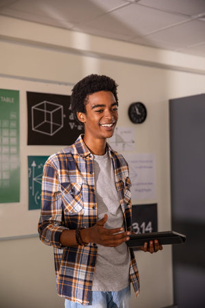 African American male teenage student gesturing and holding tablet at whiteboard in math classroom....