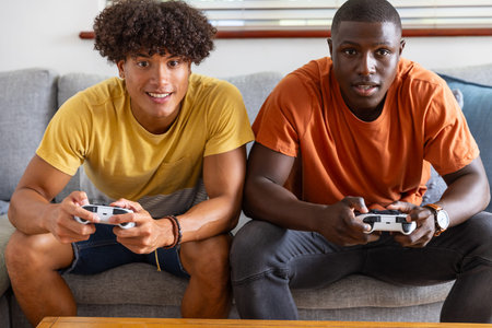Diverse male friends leaning in holding controllers on gray couch at home with blinds, coffee table....