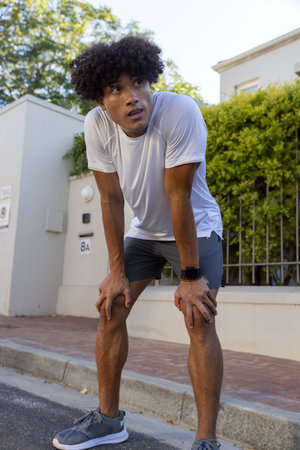 African American man in sportswear leaning forward catching breath on sidewalk checking smartwatch....