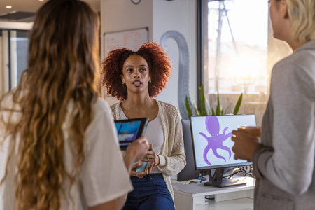 Diverse female coworkers collaborating around desk in office using tablet coffee cup and monitor....