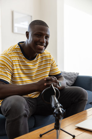 African American man leaning toward microphone beside open laptop recording podcast in living room....