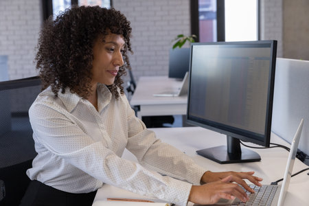 African American woman typing on laptop with monitor showing spreadsheet in office, copy space....