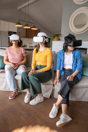 Diverse female friends testing virtual reality headsets while sitting on sofa in modern living room....