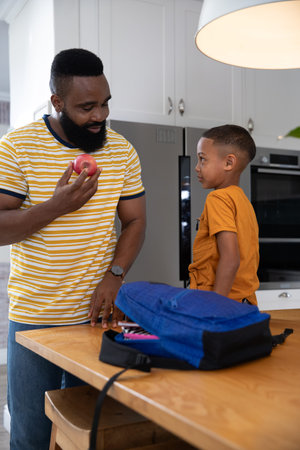 African American father and son standing at home kitchen island holding red apple and blue backpack....