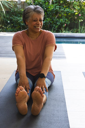 Senior African American woman stretching forward on yoga mat on deck beside pool wearing sportswear....